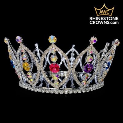 The Belle-3"x5" Fully Round-RAINBOW crown, featuring silver rhinestones and large pink, yellow, and purple gems, is shown on a black background. Ready to ship from stock at AWC RhinestoneCrowns.com, with more in production. Gold crown logo top right.