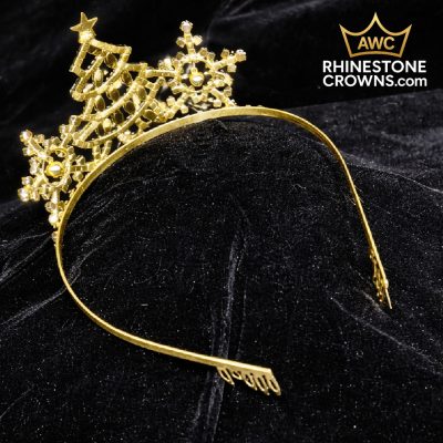 The Jingle All The Way Series-3" Wide Span Tiara is a gold rhinestone crown headband with Christmas tree and snowflake designs, perfect for quinceañeras or pageants. Displayed on black velvet. AWC RhinestoneCrowns.com logo in the top right.